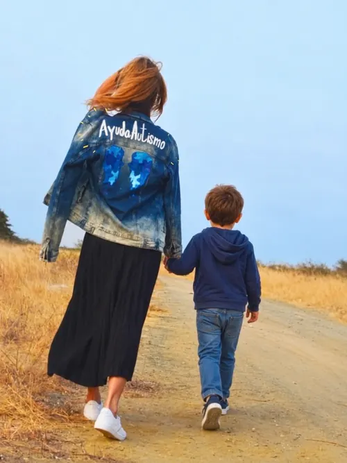 Paola Carlton camina de la mano de un niño por un sendero, con su chaqueta de AyudaAutismo